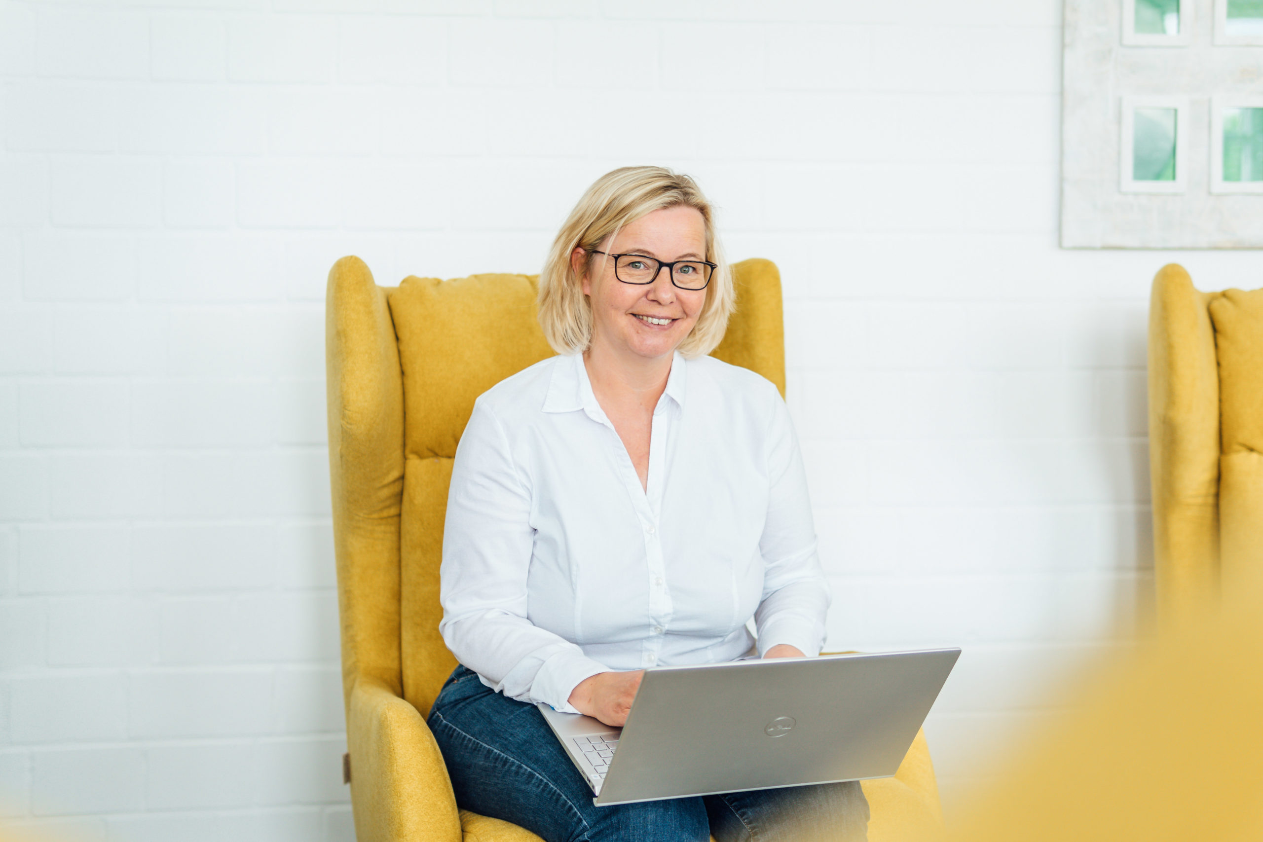 Nicole Döhrmann steht an einem Schreibtisch mit einem Laptop in der Hand. Sie trägt formelle Kleidung (Bluse und Blazer) und guckt freundlich in die Kamera